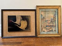 Two framed artworks side by side on floor; left artwork is lacquerware painting of two cranes, right artwork is Asian cork carving scene under glass in gold frame.