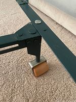 Close-up of the metal bed frame leg with a small square wood wheel attached, showing the construction and materials of the frame's support.