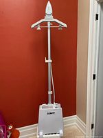 Front view of a Conair clothes steamer standing fully assembled with vertical pole and hanger against a red wall.