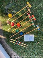 Six wooden croquet mallets with colored bands on heads and permanent colored croquet balls laid out on grass with stakes and wickets.