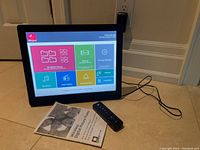 Front view of the Pixstar digital photo frame powered on showing colorful menu options, accompanied by remote control and user manual on floor.