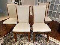 Front view of four upholstered dining chairs with cream-beige fabric and medium brown wooden legs on a patterned rug.