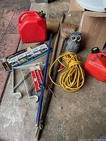 Photo showing lot items arranged on pavement with gas cans, brooms, extension cord, hooks, owl, and other items visible.