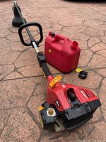 Side view of red Homelite 26SS gas trimmer and red gas can on stone pavement.