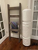 Wooden blanket ladder leaned against wall with crocheted blanket draped over a rung and Ikea standing lamp with white paper shade beside it.