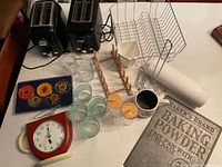 Full view of lot contents showing two black Oster toasters, glass cups, wooden dish rack, metal paper towel holder with paper towel roll, vintage metal sign, clock, and vintage Olympic coasters.