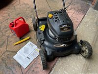 Overview of MTD black gas-powered lawnmower with red plastic gas can and owner's manual placed beside it on a concrete patio.
