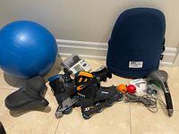 Blue 18 inch exercise ball, ObusForme back support, percussion massager with manual and accessories, two squish balls, men's size 8 Bauer roller blades with crack visible on boot, white digital blood pressure monitor, wide bike seat, and mini bike bag.