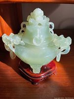 Front view of pale green intricately carved stone urn on dark wood pedestal under sunlight.