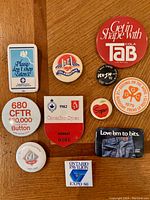Nine vintage pins laid out on wooden surface. Pins vary in design including radio station 680 CFTR, Canadian Open 1982 hockey event, Levi's denim promotion, and Expo 86 Ontario Pavilion.