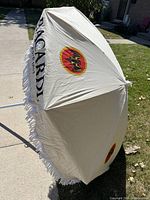 Side view of beige Bacardi patio umbrella showing the fabric canopy with Bacardi logos and white fringe trim along the edge.