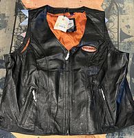 Front view of the black leather vest showing zipper closure, Harley Davidson patch on chest, and two zippered side pockets.