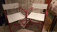 Two red metal framed director chairs with white fabric seats and backs side by side on carpeted floor.
