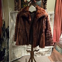 Full front view of the brown vintage fur coat hanging on wooden stand against a fabric backdrop.