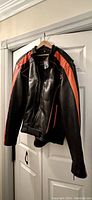 Black leather motorcycle jacket with orange stripes, displayed hanging on a door.