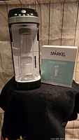 Sparkel carbonation machine with attached clear bottle and a box of carbonator packets on table.