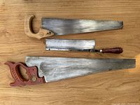Photo showing three assorted hand saws on a wooden surface: large with reddish-brown handle; medium with light wooden handle stamped 'K W Made in Germany'; small with dark wooden handle and metal blade cover.