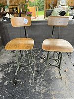 Two industrial style wooden and metal swivel bar stools with adjustable backrests shown from the front against a black wicker patio sofa, placed on a stone patio floor.