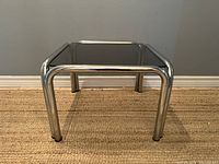 Front view of square chrome frame and smoked glass top on woven rug against grey wall