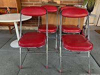 Four red vinyl and chrome folding diner-style chairs arranged together showing padded seats and backs, sitting on a tiled floor with wooden furniture in background.