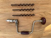Vintage hand drill ratchet brace with wooden handle and metal crank, plus three auger bits arranged above it on a wooden surface.