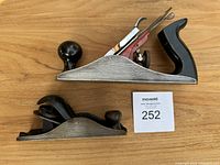 Photo of two vintage hand planers, a large Stanley planer with black wooden handles and a smaller Bailey planer, both metal-bodied.