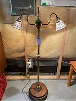 Full view of the antique two-bulb metal floor lamp standing on concrete floor against framed poster background. Shows overall design and proportions.