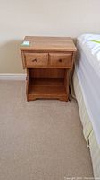 Front view of wooden bed side table with two drawers and lower shelf, sitting beside a bed on carpeted floor.