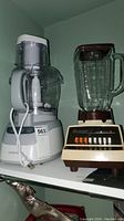 White and grey Black & Decker food processor next to Osterizer blender on white shelf.