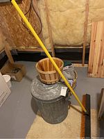 Push broom leaning on galvanized metal garbage can with lid and wooden basket on top, with towels inside basket on carpeted floor in basement