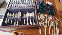 Full set of assorted silver-plated cutlery displayed in an open wooden canteen with several utensils laid out beside it on a table.