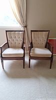 Front view of two matching sitting chairs with beige button-tufted upholstery and wooden frames in a light room.