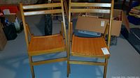 Two folding wooden chairs side by side on a blue floor with boxes and furniture in background