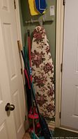 Floral patterned ironing board hanging on wall with various cleaning tools leaning beside it.