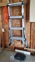 Folded aluminum telescopic articulating ladder leaning against plywood wall showing ladder structure and rubber feet.