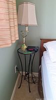 Photo shows circular metal side table with clear glass top, Curtis LED clock radio on top, and beige lamp beside bed