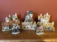 Seven miniature ceramic cottages from the 1990s David Winter collection arranged on wooden surface against red wall. Shows variety of roof styles, chimneys, and painted details.