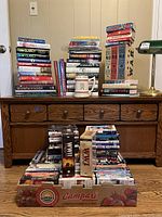 Books and DVDs stacked on and around wooden desk; various titles visible including Tom Clancy novels and military history books; DVDs stacked including documentary collections in collectible boxes.