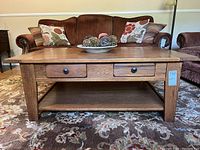 Direct front view of oak coffee table placed on patterned carpet with brown sofa set background. Two drawers visible with round black knobs.