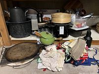 Photo showing an assortment of kitchen items including a black crockpot with steamer basket, metal cookware such as a grater and speckled pot, a green small pot with lid, wooden cutting board, and a stack of cloth linens in various colors and patterns.