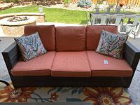 Front view of the outdoor sofa showing three orange seat cushions and two multicolor leaf pattern pillows on dark plastic wicker frame.