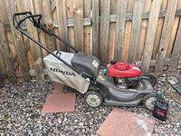 Full side view of the Honda lawn mower showing the mower with fabric grass collection bag, red engine, black deck, and wheels, on gravel with a wood fence background.