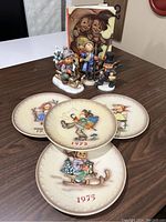 Photo of four Hummel collector plates dated 1973 and 1975, and three Hummel figurines depicting children holding lantern, sledding, and dressed in winter clothes.