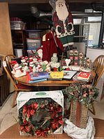 Full view of table with assorted Santa and snowman plush figurines, holiday books, cards, and a large hanging Santa decoration visible above. Two decorated wreaths and a tall decorative gift-box centerpiece in foreground.