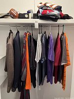 Wide view of the men's clothing lot including several suit coats, sport T-shirts hanging on a bar, folded T-shirts on a shelf, and shoes visible on top shelf.