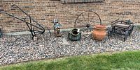 Full view of all items in the lot arranged on rock bed against brick wall including hand plow, wagon wheels, wooden bear, pottery, kettle, and metal cart.