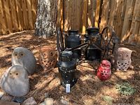 Overall lot showing owl decor in various materials including stone, terracotta, and metal, several black metal milk jugs, and antique wagon wheels in background.
