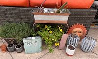 Wide view of outdoor patio decor showing multiple plant containers, a metal planter with cactus, yellow flowering plants, decorative sun face, and woven basket planters on patio floor.
