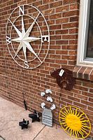 Wide view showing multiple metal outdoor decor pieces against brick wall including compass rose, candle holders, flower stake, metal sign, and yellow sun face.