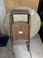Used Samsonite beige round card table leaning against wall with four folding chairs stacked in front showing wear on seats